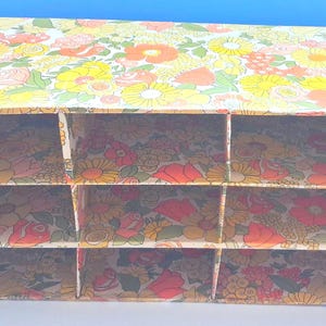 K&ouml;nnte beinhalten: Ein rechteckiger Schreibtisch-Organizer aus Pappe mit einem lebendigen Blumenmuster in Gelb-, Orange-, Rot- und Gr&uuml;nt&ouml;nen. Die Einheit ist in neun gleiche F&auml;cher unterteilt, die in drei horizontalen Reihen und drei vertikalen Spalten angeordnet sind.