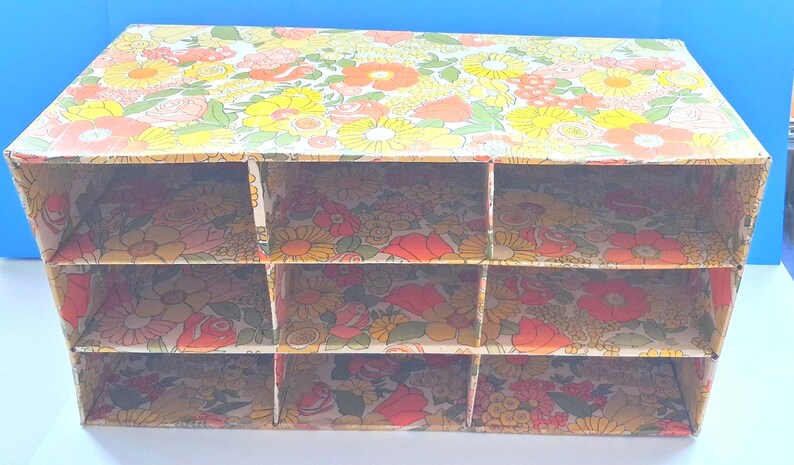 K&ouml;nnte beinhalten: Ein rechteckiger Schreibtisch-Organizer aus Pappe mit einem lebendigen Blumenmuster in Gelb-, Orange-, Rot- und Gr&uuml;nt&ouml;nen. Die Einheit ist in neun gleiche F&auml;cher unterteilt, die in drei horizontalen Reihen und drei vertikalen Spalten angeordnet sind.