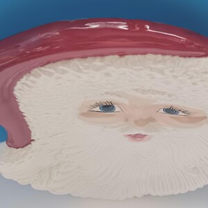 May include: A ceramic Santa Claus face with a red hat and white beard. The face has blue eyes and a pink mouth. The background is a solid blue color.