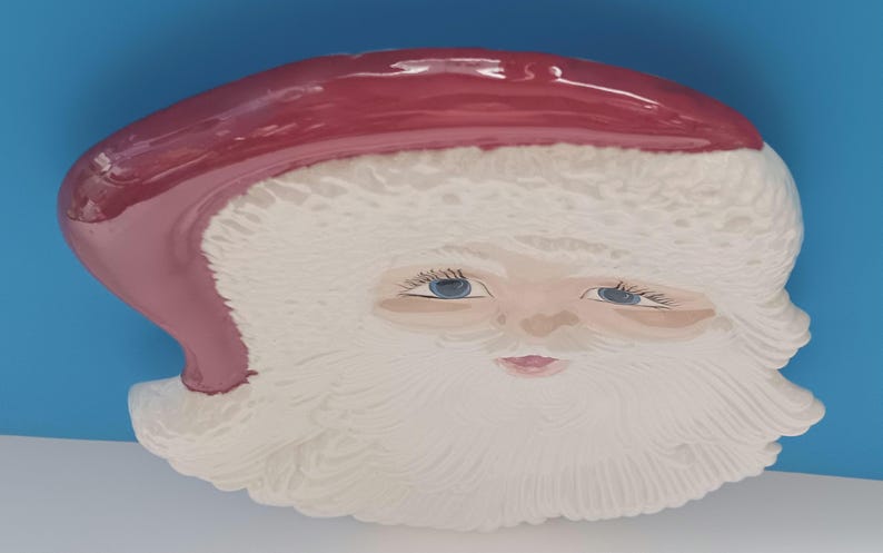 May include: A ceramic Santa Claus face with a red hat and white beard. The face has blue eyes and a pink mouth. The background is a solid blue color.
