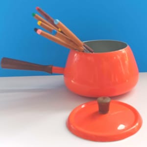 May include: An orange fondue pot with a wooden handle and lid. The pot contains several fondue forks with colored tips. The lid is next to the pot. The background is blue and the surface is white.