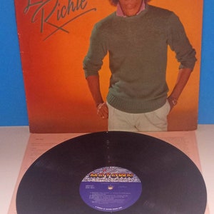 May include: Album cover for Lionel Richie. The cover features a portrait of Lionel Richie in a green sweater and white pants against an orange background. A vinyl record with a purple and gold label is in the foreground.
