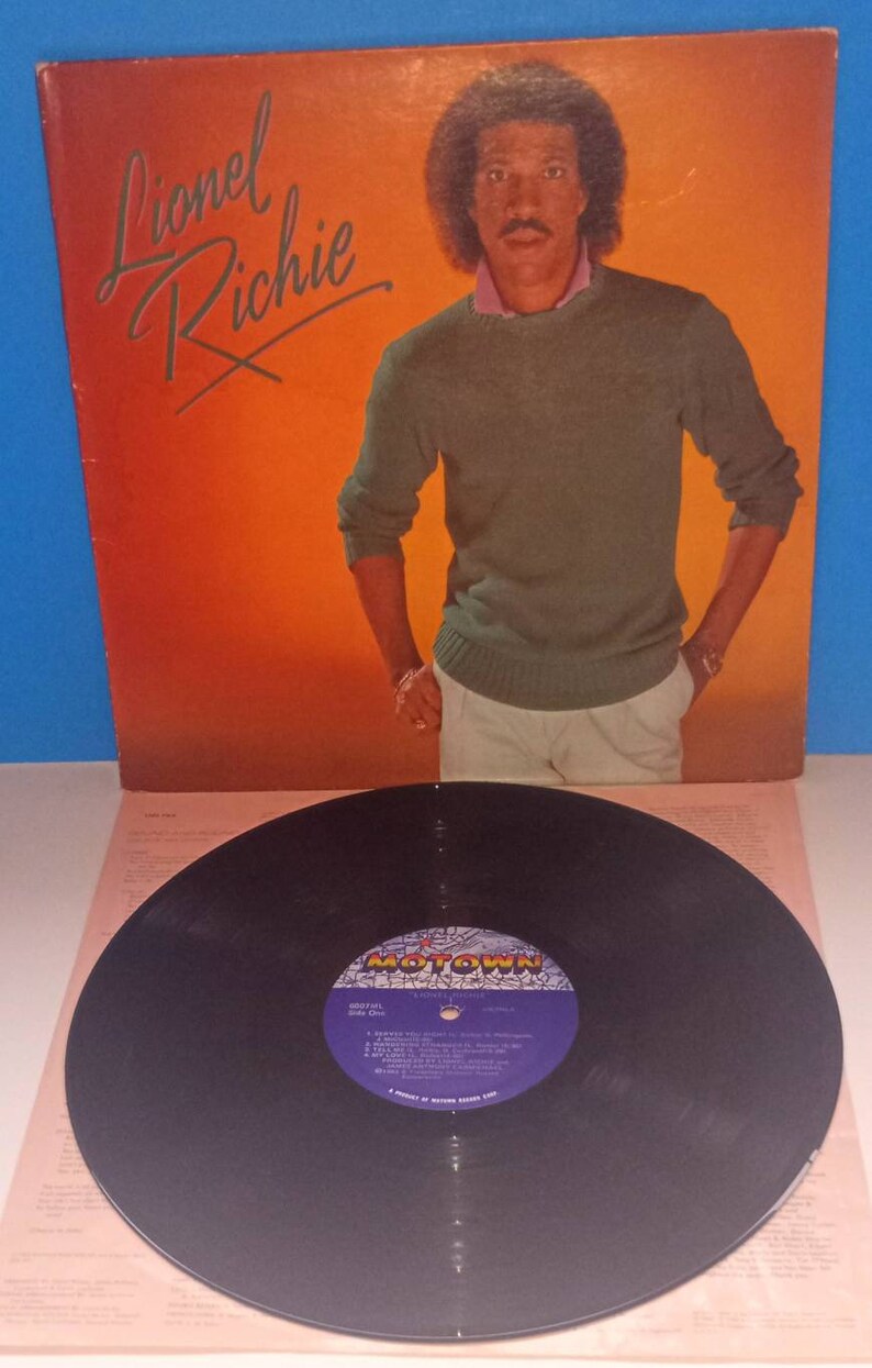May include: Album cover for Lionel Richie. The cover features a portrait of Lionel Richie in a green sweater and white pants against an orange background. A vinyl record with a purple and gold label is in the foreground.