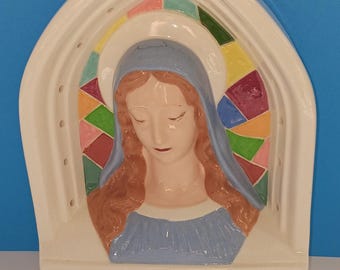 May include: A ceramic wall hanging depicting a religious figure with a blue veil and long red hair. The figure is set against a stained-glass window design with various colors. The piece has a white arched frame and a light blue base with small holes.