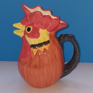May include: A ceramic pitcher shaped like a rooster. The pitcher features a red comb and wattle, yellow beak and neck, and a brown body with a black handle. The pitcher is a decorative kitchenware item.