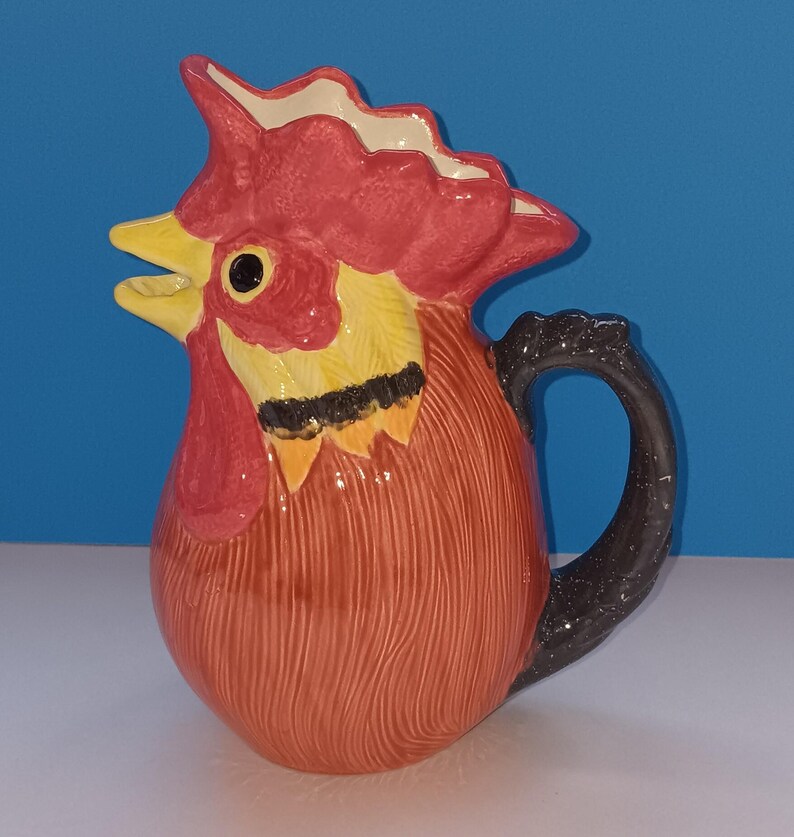 May include: A ceramic pitcher shaped like a rooster. The pitcher features a red comb and wattle, yellow beak and neck, and a brown body with a black handle. The pitcher is a decorative kitchenware item.