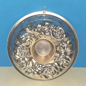 Puede incluir: Un molde de reposter&iacute;a circular de metal plateado con un patr&oacute;n floral en relieve de rosas alrededor de un n&uacute;cleo central hueco. El molde incluye un peque&ntilde;o lazo de metal integrado en el borde superior para colgarlo, con un acabado liso.