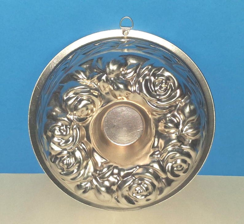 Puede incluir: Un molde de reposter&iacute;a circular de metal plateado con un patr&oacute;n floral en relieve de rosas alrededor de un n&uacute;cleo central hueco. El molde incluye un peque&ntilde;o lazo de metal integrado en el borde superior para colgarlo, con un acabado liso.