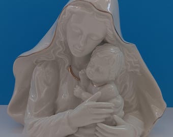 May include: A white porcelain sculpture of a mother and child. The mother figure is draped in a flowing veil with gold trim, cradling the child. The sculpture has a glossy finish and is set against a blue background.