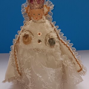 May include: A porcelain doll figurine of a young girl wearing a white dress with lace trim and a crown. The doll has a pink and gold crown with glitter and is holding a small bouquet of flowers.