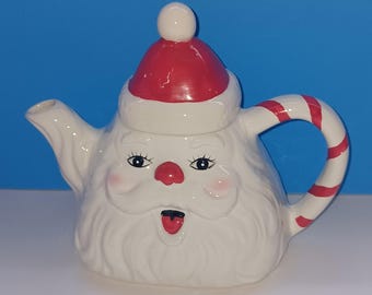 May include: A white ceramic teapot shaped like Santa Claus, featuring a red hat, nose, and candy cane-striped handle. The teapot has a smiling face with rosy cheeks and a small red tongue. A festive holiday item.