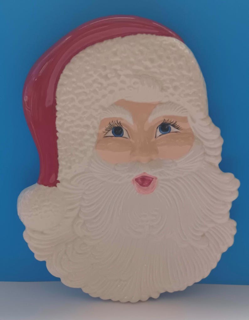 May include: A Santa Claus face-shaped decorative item. The item features a red hat, a white beard, and a rosy face with blue eyes. The item is likely made of ceramic or a similar material, and is designed for holiday decor.