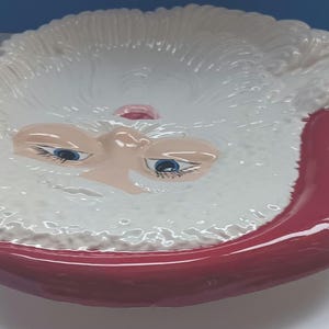 May include: A ceramic Santa Claus face-shaped dish. The dish features a white beard and eyebrows, a pink nose, and blue eyes. The rim of the dish and the mouth are a deep red color. This decorative dish is perfect for holiday decor.