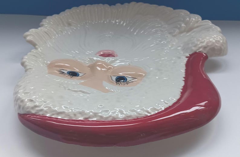 May include: A ceramic Santa Claus face-shaped dish. The dish features a white beard and eyebrows, a pink nose, and blue eyes. The rim of the dish and the mouth are a deep red color. This decorative dish is perfect for holiday decor.