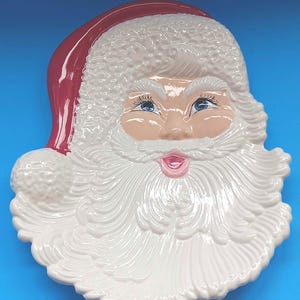 May include: A ceramic Santa Claus face-shaped serving platter. The platter features a red hat, rosy cheeks, blue eyes, and a white beard. The plate is a festive holiday decoration.