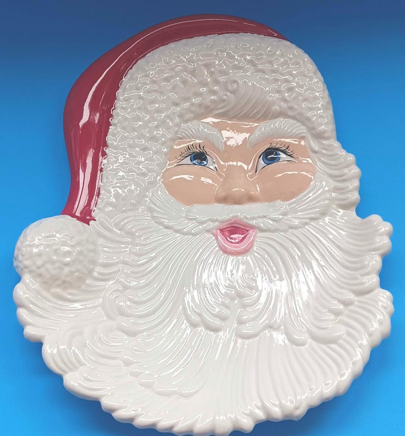 May include: A ceramic Santa Claus face-shaped serving platter. The platter features a red hat, rosy cheeks, blue eyes, and a white beard. The plate is a festive holiday decoration.