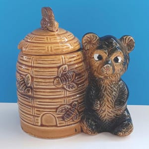 May include: A ceramic honey jar and bear figurine. The jar is shaped like a beehive with a lid and bee details. The bear is brown and black with white eyes. The jar and bear are tan and brown.