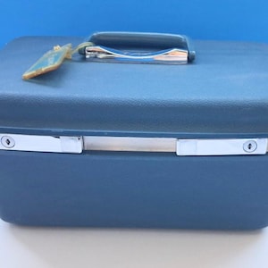 May include: A vintage navy blue travel case with a silver handle and latches. The case has a rectangular shape with rounded corners and a matching navy blue handle. A small tag hangs from the handle.