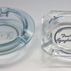 May include: A round, light blue glass ashtray with the text RAPA and a square, clear glass ashtray featuring the text Fontana's Spaghetti House, Cheektowaga: 822-7918, and 4446 printed in the center. Both items feature notches along the rim.