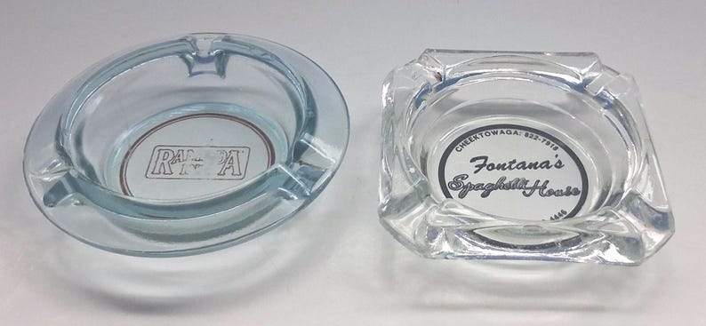 May include: A round, light blue glass ashtray with the text RAPA and a square, clear glass ashtray featuring the text Fontana's Spaghetti House, Cheektowaga: 822-7918, and 4446 printed in the center. Both items feature notches along the rim.