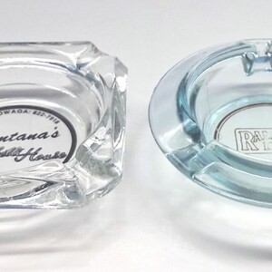 May include: Two clear glass ashtrays. The square ashtray features the black text Fontana's Spaghetti House and Cheektowaga 822-7918. The round ashtray has a light blue tint and features a rectangular logo with the letters R and A inside.