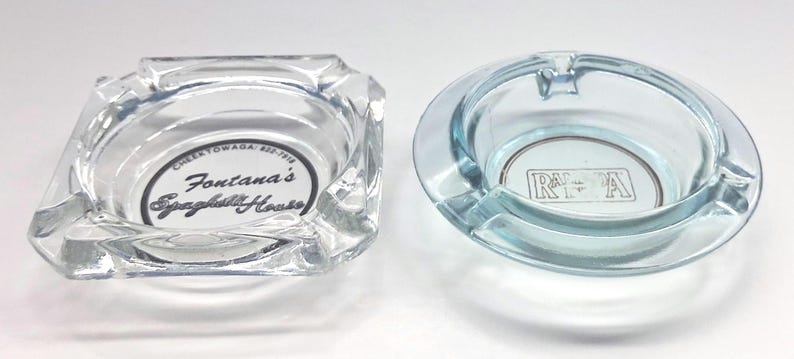 May include: Two clear glass ashtrays. The square ashtray features the black text Fontana's Spaghetti House and Cheektowaga 822-7918. The round ashtray has a light blue tint and features a rectangular logo with the letters R and A inside.