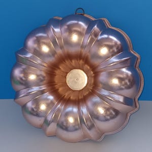 May include: A vintage copper-colored bundt cake pan with a decorative fluted design. The pan has a central tube and a hanging loop. The metallic surface reflects light, creating highlights and shadows. The pan is designed for baking cakes.