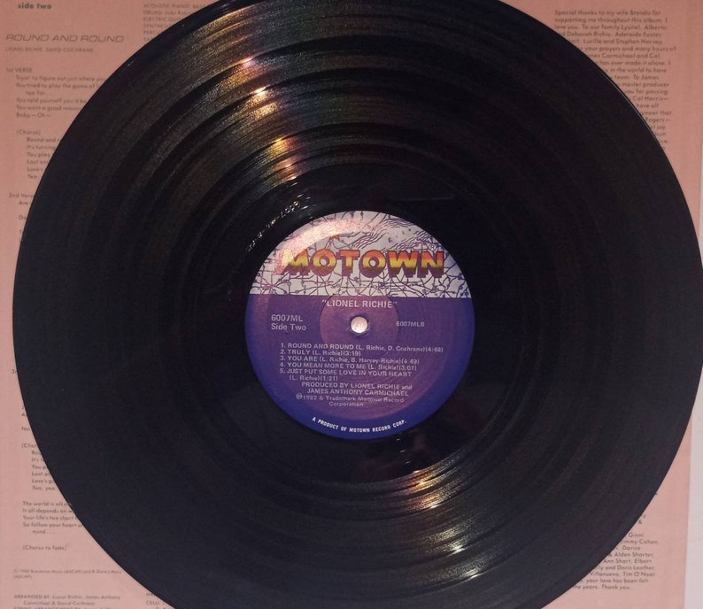 May include: A black vinyl record with the label "MOTOWN" and the artist "LIONEL RICHIE." The record is labeled "Side Two" and lists song titles including "Round and Round" and "Truly." The record is on a pink background.