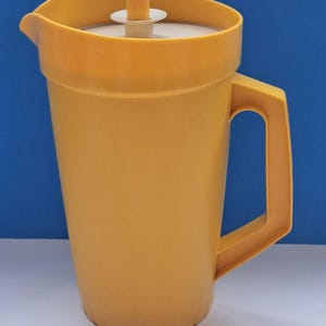 May include: A yellow plastic pitcher with a matching handle and lid. The pitcher has a spout for easy pouring and a white interior. The lid features a small, round handle. The pitcher is designed for holding liquids.