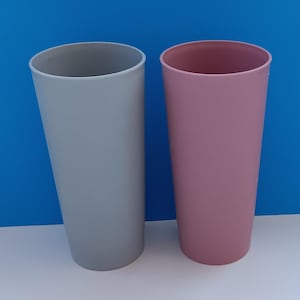 Puede incluir: Dos vasos altos de plástico, uno gris y otro rosa. Los vasos están vacíos y están sobre una superficie blanca.