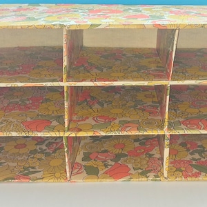 K&ouml;nnte beinhalten: Ein rechteckiger Aufbewahrungsorganizer mit neun offenen F&auml;chern, die in drei Reihen und drei Spalten angeordnet sind. Die gesamten Au&szlig;en- und Innenfl&auml;chen sind mit einem Blumenmuster aus gelben, orangefarbenen und rosafarbenen Bl&uuml;ten auf cremefarbenem Grund bedeckt.