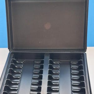 May include: An open, black plastic carrying case with a handle. The interior is divided into compartments, with slots and ridges designed to hold items. The case is empty and sits on a white surface against a blue background.