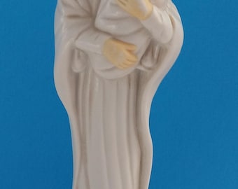 Op de afbeelding: Een keramieken beeldje van Maria die baby Jezus vasthoudt, beiden in witte gewaden. Maria heeft bruin haar en een serene uitdrukking. De baby is in een witte deken gewikkeld. De achtergrond is effen blauw.