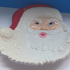 May include: A ceramic Santa Claus face-shaped serving dish. The dish features a red hat, a white beard, and a rosy face with blue eyes and a pink mouth. The dish is white with a glossy finish, perfect for holiday gatherings.