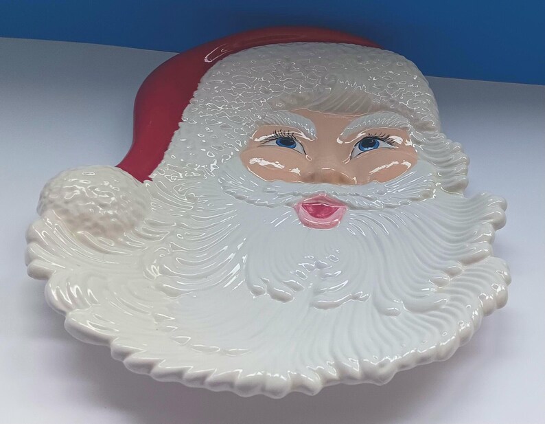 May include: A ceramic Santa Claus face-shaped serving dish. The dish features a red hat, a white beard, and a rosy face with blue eyes and a pink mouth. The dish is white with a glossy finish, perfect for holiday gatherings.