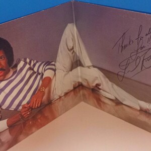 May include: Album cover featuring a man in a striped shirt and white pants. The man is lying down, with a signature and the words "Thanks for all you Love!!" written on the cover. The album is folded.