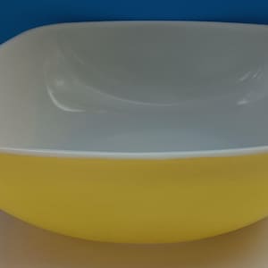 Puede incluir: Un cuenco de estilo retro con exterior amarillo e interior blanco. El cuenco tiene una forma asimétrica inusual con bordes redondeados y un acabado liso y brillante. Una pieza clásica para servir o exhibir, que recuerda el diseño de mediados de siglo.