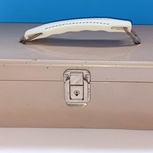 May include: A vintage metal toolbox in a muted rose-gold color. It features a white handle with a stitched design and a silver clasp closure. The box is rectangular and appears to be made of durable metal.