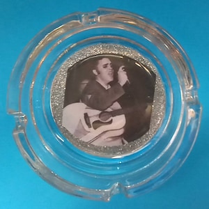 May include: Circular clear glass ashtray. The centre has a black and white image of a musician with a guitar, framed by silver glitter. The ashtray has three cigarette rests. The ashtray is made of clear glass.