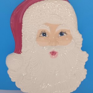 May include: A ceramic Santa Claus face with a white beard and hat, and a pink hat. The Santa face has blue eyes and a pink mouth. The background is a solid blue.