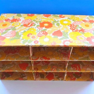 K&ouml;nnte beinhalten: Ein rechteckiger Aufbewahrungsorganizer aus Pappe mit einem lebendigen Blumenmuster in Gelb-, Orange-, Rot- und Gr&uuml;nt&ouml;nen. Die Einheit ist in neun einzelne F&auml;cher unterteilt, die in drei Reihen und drei Spalten angeordnet sind, mit einer offenen Vorderseite.