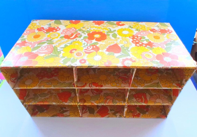 K&ouml;nnte beinhalten: Ein rechteckiger Aufbewahrungsorganizer aus Pappe mit einem lebendigen Blumenmuster in Gelb-, Orange-, Rot- und Gr&uuml;nt&ouml;nen. Die Einheit ist in neun einzelne F&auml;cher unterteilt, die in drei Reihen und drei Spalten angeordnet sind, mit einer offenen Vorderseite.