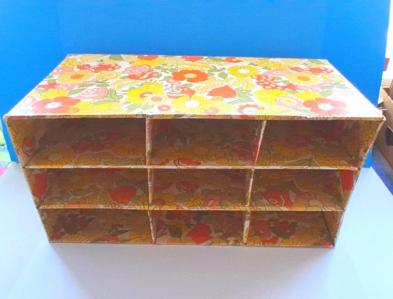 K&ouml;nnte beinhalten: Ein rechteckiger Schreibtisch-Organizer aus Pappe mit neun offenen F&auml;chern, die in drei Reihen und drei Spalten angeordnet sind. Die gesamten Au&szlig;en- und Innenfl&auml;chen sind mit einem lebendigen Blumenmuster in Gelb-, Orange- und Rott&ouml;nen bedeckt.