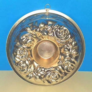 Puede incluir: Un molde para gelatina redondo de color dorado met&aacute;lico con un patr&oacute;n floral en relieve de rosas alrededor del centro. El molde tiene una base circular lisa en el medio e incluye una peque&ntilde;a anilla met&aacute;lica en el borde superior para colgar.