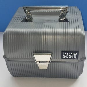 May include: A gray, ribbed, hard-shell storage case with a silver-toned metal handle and a central metal latch. The front features a small, rectangular black label with the white text SASSABY printed in a stylized font on the lower right side.