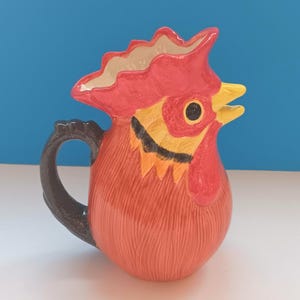 May include: A ceramic pitcher shaped like a rooster's head. The pitcher is primarily orange with red, yellow, black, and white accents. The handle is black. The rooster's comb and wattle are red, and the beak is yellow.