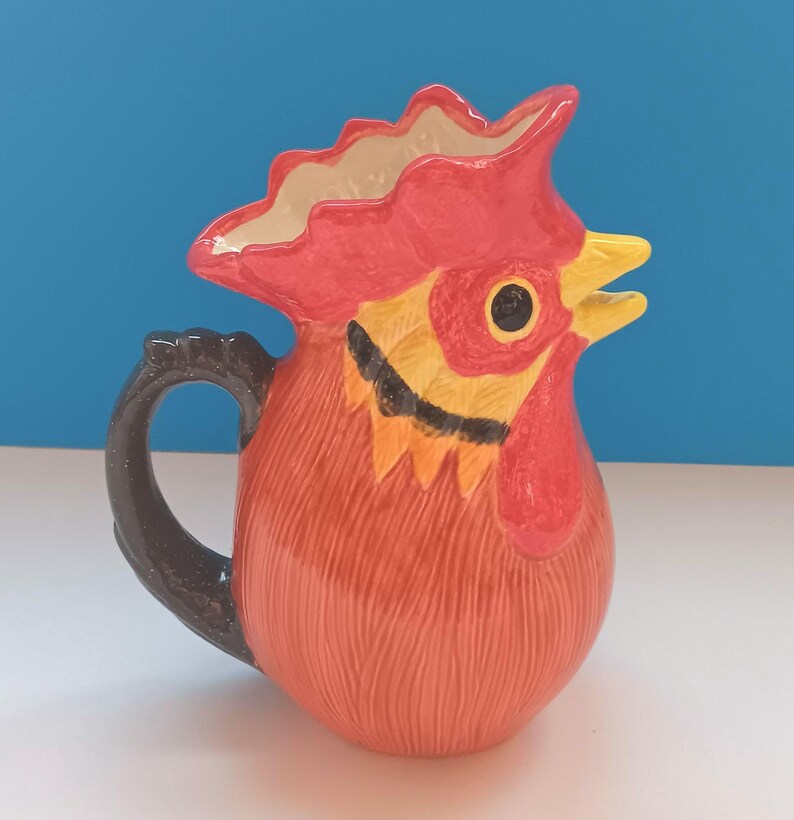 May include: A ceramic pitcher shaped like a rooster's head. The pitcher is primarily orange with red, yellow, black, and white accents. The handle is black. The rooster's comb and wattle are red, and the beak is yellow.