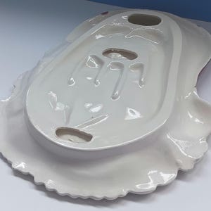 May include: A white ceramic serving dish with a scalloped edge. The dish has a unique shape with a raised center section and three oval cutouts. The underside is visible, showing the dish's form and design.