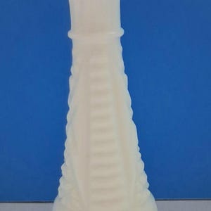 May include: A white, vintage-style vase with a textured, patterned design. The vase has a flared top and a rounded base. The body of the vase features vertical ridges and a series of horizontal lines. The vase is made of a solid, opaque material.