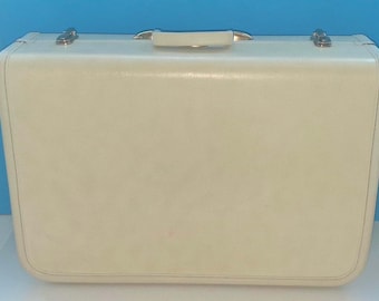 May include: A rectangular off-white suitcase with a smooth, solid-colored exterior. The luggage features a top-mounted carrying handle and two metallic silver-toned latches positioned on the upper edge for secure closure of the main compartment.